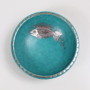 May include: A teal decorative bowl featuring a silver fish design. The bowl has a scalloped silver rim. The fish is detailed with scales and fins, and bubbles are depicted around it. The bowl's diameter is approximately 25 cm.