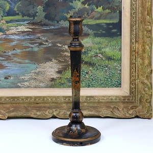 Peut inclure: Un chandelier noir antique avec des détails dorés. Le chandelier a une base ronde, une tige fine et un dessus décoratif. La tige présente un motif peint avec un bâtiment. Le chandelier est placé devant une peinture de paysage.