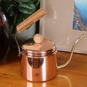 May include: A copper gooseneck kettle with a wooden handle and knob. The spout is gold-coloured. This kettle is designed for precise pouring, often used for pour-over coffee. The kettle's design is reminiscent of vintage kitchenware.