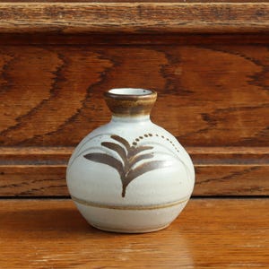 May include: A small, round ceramic vase with a cream-colored glaze. The vase features a hand-painted brown leaf motif on the front and a brown rim around the narrow opening. A thin brown horizontal line encircles the lower portion of the vase body.