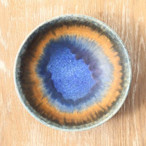 May include: A round ceramic bowl featuring a reactive glaze with a deep blue center that transitions into a vibrant orange band, surrounded by a dark brown and grey rim. The interior surface displays a smooth, glossy finish with blended color gradients.