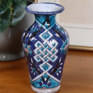 May include: A small, ceramic vase with a detailed, geometric pattern in shades of blue, turquoise, and white. The vase has a narrow neck and a flared rim. The base is a light gray color. The vase is likely handmade and suitable for small floral arrangements.