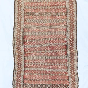 Puede incluir: Textil rectangular tejido con un patrón geométrico en tonos rojos, marrones y beige. Los bordes están bordeados con un patrón de diamantes y los extremos están deshilachados. Probablemente sea una alfombra o un tapiz.