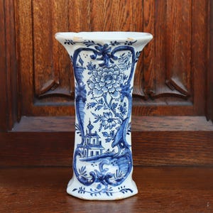 May include: A white ceramic vase featuring blue hand-painted floral patterns and a landscape scene with a building. The vase has a flared top and a curved, tapered body with ornate scrollwork framing the detailed blue illustrations.