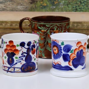 May include: Three decorative mugs. Two white mugs with blue and orange floral designs and a brown mug with green floral accents. The mugs are on a white surface.