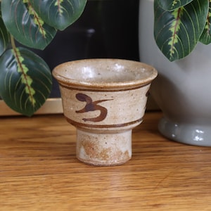 Puede incluir: Una pequeña taza de cerámica hecha a mano, con una base beige y un borde marrón oscuro y un elemento decorativo. La taza tiene una base de pedestal y se exhibe sobre una superficie de madera. La taza probablemente se usa para beber.