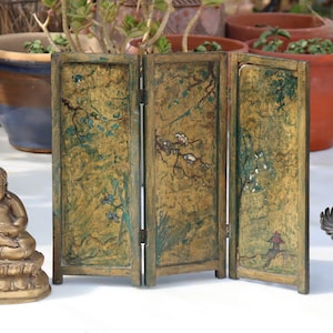 May include: A three-panel folding screen featuring a distressed gold finish with hand-painted green foliage, blue flowers, and a small red structure. The screen is constructed with hinged wooden frames and displays a weathered, textured surface.
