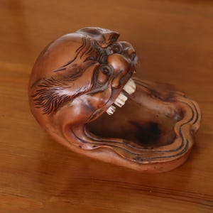 Könnte beinhalten: Eine geschnitzte Holzskulptur einer Kreatur mit offenem Mund und sichtbaren Zähnen. Die Skulptur ist braun mit dunklen Akzenten und hat eine tablettartige Basis. Das Stück ist wahrscheinlich ein Dekorationsartikel oder ein kleiner Behälter.