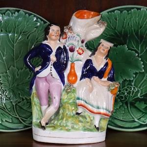 May include: A vintage ceramic pitcher featuring a man and woman in period clothing. The pitcher has a white base with colorful accents, including orange, blue, and green. The figures are set against a backdrop of stylized foliage and flowers.