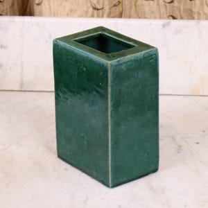 Pode incluir: Um vaso retangular de cerâmica verde esmeralda com uma abertura quadrada. O vaso tem um acabamento brilhante e é exibido em uma superfície de mármore branco. O vaso é um item decorativo.