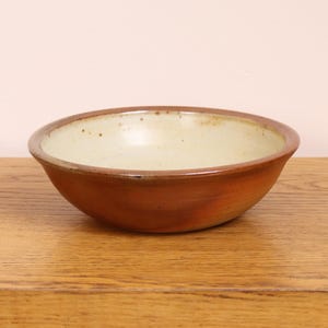 Puede incluir: Un cuenco de cerámica hecho a mano con una paleta de colores cálidos y terrosos. El cuenco presenta un interior beige claro y un exterior marrón rojizo intenso. Se encuentra sobre una superficie de madera, mostrando su diseño simple pero elegante. Ideal para servir alimentos.