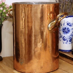 May include: A tall, cylindrical copper bucket with two brass handles. The bucket has a polished, reflective surface and a slightly aged appearance. The handles are ornate and curved. The bucket is likely used for decorative or storage purposes.