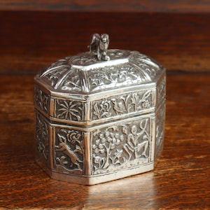 May include: A silver-coloured metal box with an octagonal shape and a removable lid. The exterior surfaces feature intricate embossed relief carvings of figures, trees, and animals. A small sculpted animal figure sits atop the centre of the lid.