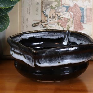 Puede incluir: Un cuenco de cerámica hecho a mano con la parte superior cuadrada y la base redondeada. El cuenco está esmaltado en negro con un esmalte blanco goteado alrededor del borde. El cuenco está sobre una superficie de madera.