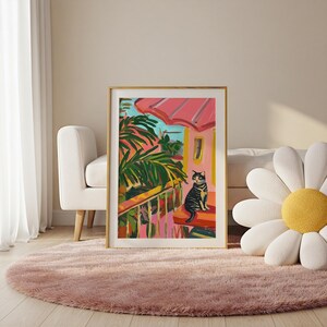 May include: A framed art print showing a vibrant painting of a cat on a porch. The artwork features a pink roof, green palm leaves, and a blue sky. The cat is black and grey. The painting is in a gold frame, adding to its aesthetic appeal.