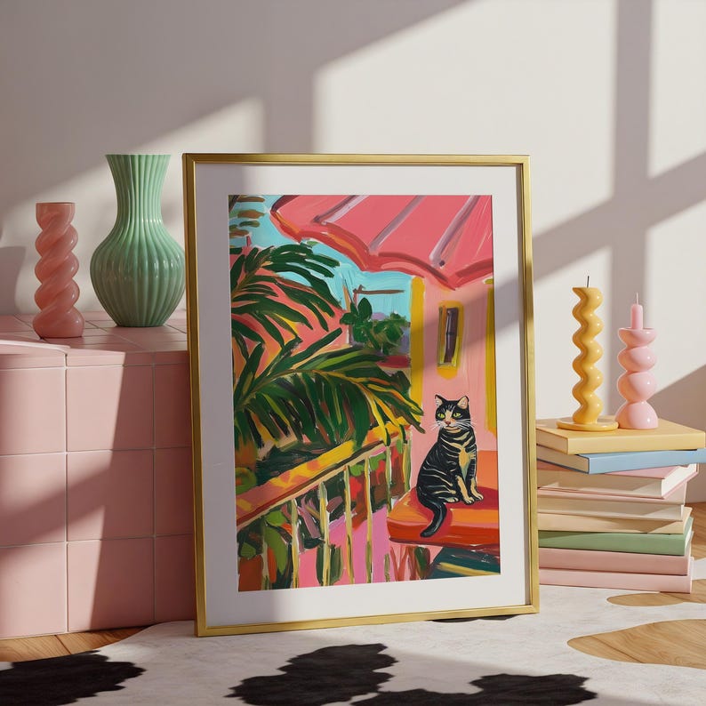 May include: A framed art print with a gold frame shows a cat on a porch. The artwork features a pink roof, green palm leaves, and a yellow railing. Decorative vases and candles are in the background.