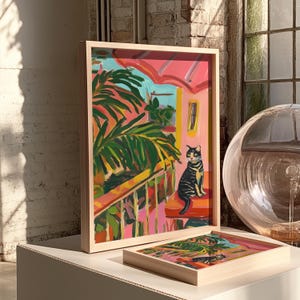 May include: A framed art print depicting a vibrant painting of a black and yellow cat on a balcony. The artwork has a pink and green tropical theme, with a light wood frame. A second, smaller print is also visible.