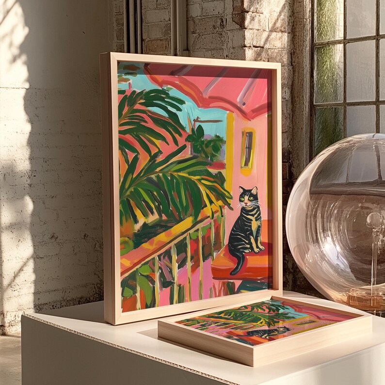 May include: A framed art print depicting a vibrant painting of a black and yellow cat on a balcony. The artwork has a pink and green tropical theme, with a light wood frame. A second, smaller print is also visible.