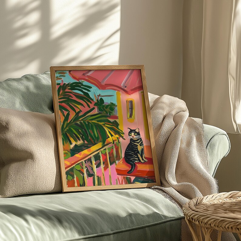May include: A framed art print showing a painting of a black and grey cat. The artwork depicts a tropical scene with palm fronds, a pink roof, and a balcony with red and white railings. The frame is made of light-coloured wood. The painting is set on a red surface.
