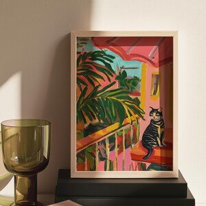 May include: A framed art print showing a vibrant outdoor scene. A black and yellow tabby cat sits on a red cushion on a balcony. The artwork features palm fronds, a pink building, and a bright blue sky.