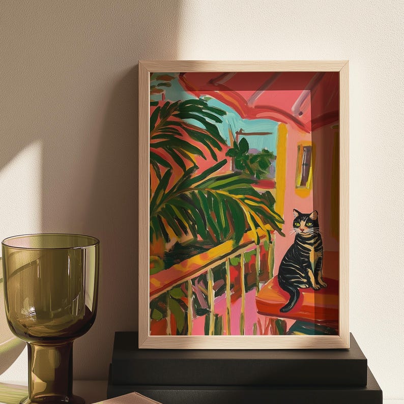 May include: A framed art print showing a vibrant outdoor scene. A black and yellow tabby cat sits on a red cushion on a balcony. The artwork features palm fronds, a pink building, and a bright blue sky.
