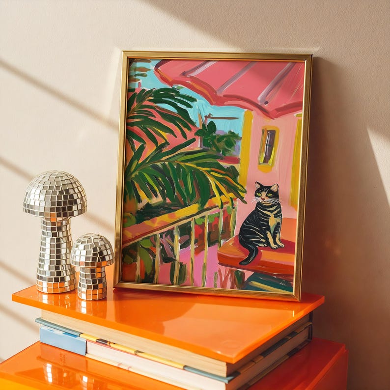 May include: A framed painting of a cat on a balcony with a tropical scene. The painting is in a gold frame and sits on a stack of orange books. Two mirrored mushroom-shaped decorative objects are on the shelf.