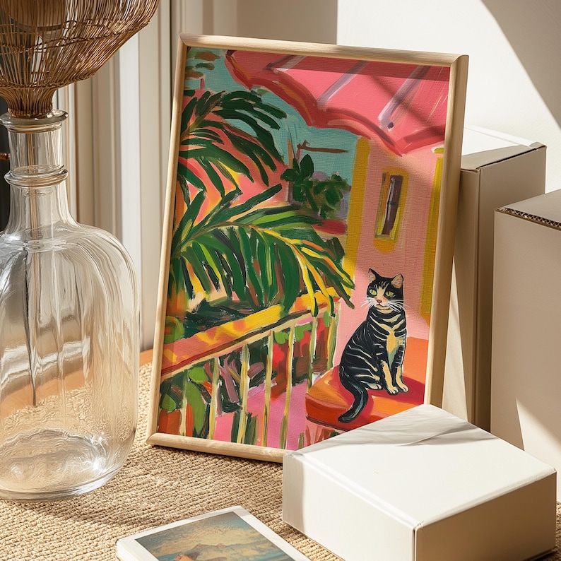 May include: A framed painting depicting a cat on a balcony with palm trees. The artwork features a black and yellow cat, a pink awning, and a yellow railing. The painting is in a light wood frame.
