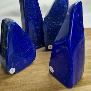 Peut inclure: Quatre pierres de lapis-lazuli polies de tailles variées, chacune d'un bleu profond et veinée naturellement. Les pierres sont exposées sur une surface en bois, mettant en valeur leurs formes et textures uniques. Une belle collection.