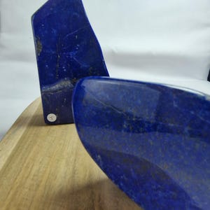 Può includere: Due pietre di lapislazzuli blu intenso lucidate con venature dorate naturali. Le pietre sono esposte su una superficie di legno su sfondo bianco. Una pietra è in posizione verticale, l'altra è angolata.