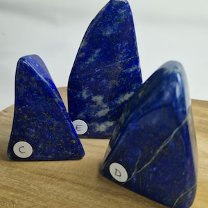 Peut inclure: Trois sculptures en pierre de lapis-lazuli polies. Chaque pierre est d'un bleu profond avec des veines blanches et dorées. Les pierres sont de forme triangulaire et reposent sur une surface en bois.