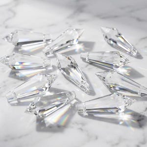 May include: A scattering of clear, faceted crystal pendants on a white surface. Each pendant has a pointed end and a rectangular base. The crystals reflect light, creating a sparkling effect. These pendants are likely for decorative purposes.