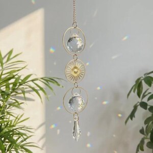 May include: A hanging suncatcher with a gold chain and circular frames. Two clear crystal balls and a crystal teardrop dangle from the chain. A gold sunburst design is centered between the crystal balls. The suncatcher is suspended in front of a light background.