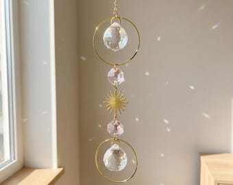 May include: A hanging suncatcher with a gold chain and accents. The suncatcher features two clear crystal balls, two pink crystal gems, and a gold sunburst charm. The suncatcher is suspended in front of a window.