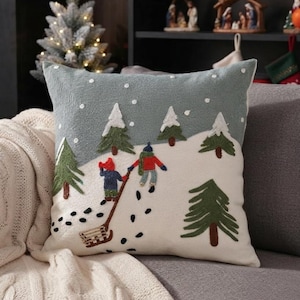 May include: A square throw pillow featuring an embroidered winter scene with a light gray sky, white snow, green pine trees, and two figures in red and blue winter clothing pulling a wooden sled across a snowy landscape with white dots.