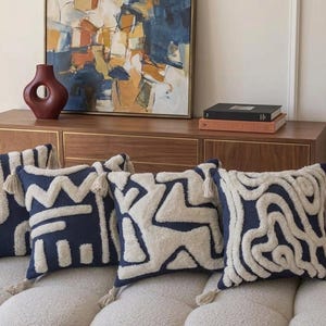 Könnte beinhalten: Vier marineblaue Zierkissen mit cremefarbenen geometrischen Mustern und Quasten. Die Kissen liegen auf einem cremefarbenen Sofa. Im Hintergrund ein Holzschrank mit einem Gemälde und einer weinroten Vase.