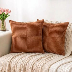 Puede incluir: Dos cojines cuadrados de pana en un cálido tono marrón terroso. Cada cojín presenta un diseño de patchwork con secciones acanaladas verticales y horizontales. Los cojines descansan sobre una manta de punto trenzado color crema.