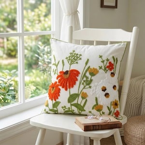 Peut inclure: Un coussin carré blanc avec des fleurs brodées orange, jaunes et blanches et des tiges et feuilles vertes. Le coussin a une bordure verte et est posé sur une chaise blanche. Un livre et des lunettes reposent sur la chaise.