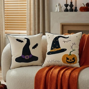 May include: Two square scatter cushions with cream-coloured fabric and black piping. One cushion features a black witch hat with a purple band and two black bats. The other cushion displays a black witch hat with a yellow band and a jack-o'-lantern.