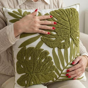 Puede incluir: Cojín cuadrado blanco con un diseño de hojas de monstera verdes. Las hojas son texturizadas y en relieve sobre el fondo blanco. El cojín tiene un borde verde y es un elemento decorativo para el hogar.