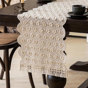 May include: An off-white table runner featuring an intricate floral lace pattern with scalloped edges and a fringe trim at the bottom. The fabric displays a repeating design of small, raised flower motifs and eyelet cutouts throughout the length.