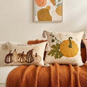 Pode incluir: Dois almofadas decorativas na cor creme com bordado texturizado. A almofada retangular apresenta cinco abóboras e a palavra "autumn" em marrom. A almofada quadrada exibe uma grande abóbora amarela com folhas e vinhas verdes.