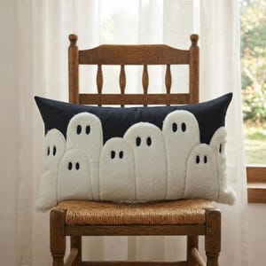 May include: A rectangular throw cushion featuring a dark charcoal background with a series of white, fuzzy, ghost-shaped appliqués. Each ghost figure has two simple, dark, oval-shaped eyes, creating a repeating pattern across the front surface.