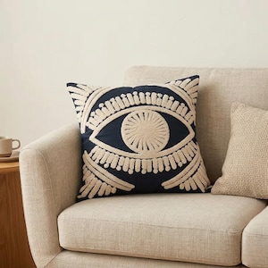 May include: A navy blue square throw pillow with an embroidered eye design in beige. The pillow is placed on a beige sofa, with another beige pillow visible. The eye design features a central circle and surrounding details.