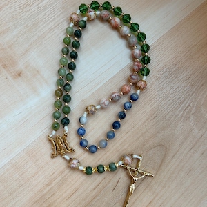 Puede incluir: Un rosario con cuentas de varios colores, incluyendo verde, azul y melocotón, conectadas por eslabones de metal dorado. El rosario presenta un crucifijo dorado y una pieza central decorativa con la letra "M".