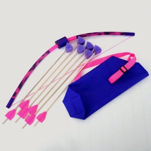 May include: A toy archery set featuring a pink and purple bow with matching arrows and a blue quiver. The bow has a patterned design, and the arrows have pink fletching. The quiver has a pink strap.