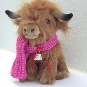 K&ouml;nnte beinhalten: Ein Stofftier, das eine Highland-Kuh darstellt, mit zotteligem, braunem Fell, einem rosa gestrickten Schal und einer silbernen Glocke. Das Spielzeug hat kleine schwarze H&ouml;rner und ein hellbraunes Gesicht. Die Kuh sitzt aufrecht.