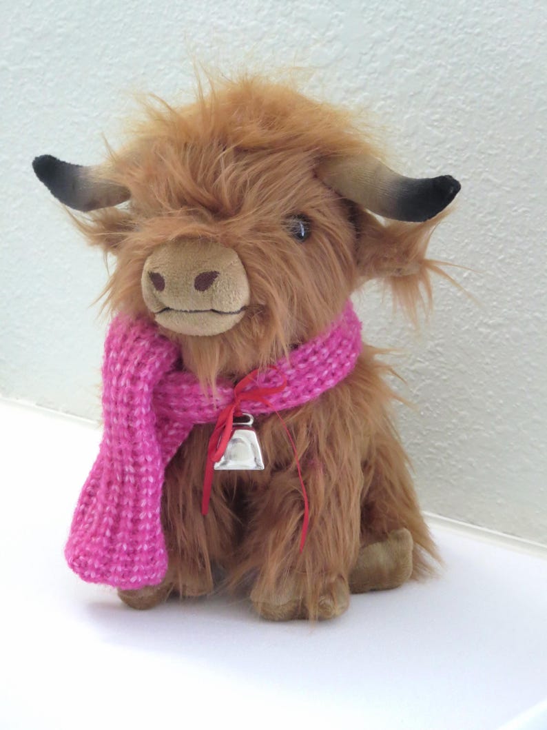 K&ouml;nnte beinhalten: Ein Stofftier, das eine Highland-Kuh darstellt, mit zotteligem, braunem Fell, einem rosa gestrickten Schal und einer silbernen Glocke. Das Spielzeug hat kleine schwarze H&ouml;rner und ein hellbraunes Gesicht. Die Kuh sitzt aufrecht.