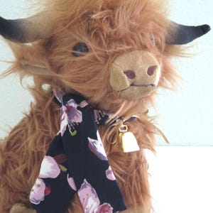 K&ouml;nnte beinhalten: Ein Pl&uuml;sch-Highland-Kuh mit zotteligem braunem Fell, schwarzen H&ouml;rnern und einer beigen Schnauze. Sie tr&auml;gt einen schwarzen Schal mit rosa Blumenmustern und eine kleine goldene Glocke. Das Spielzeug ist einer Highland-Kuh nachempfunden.