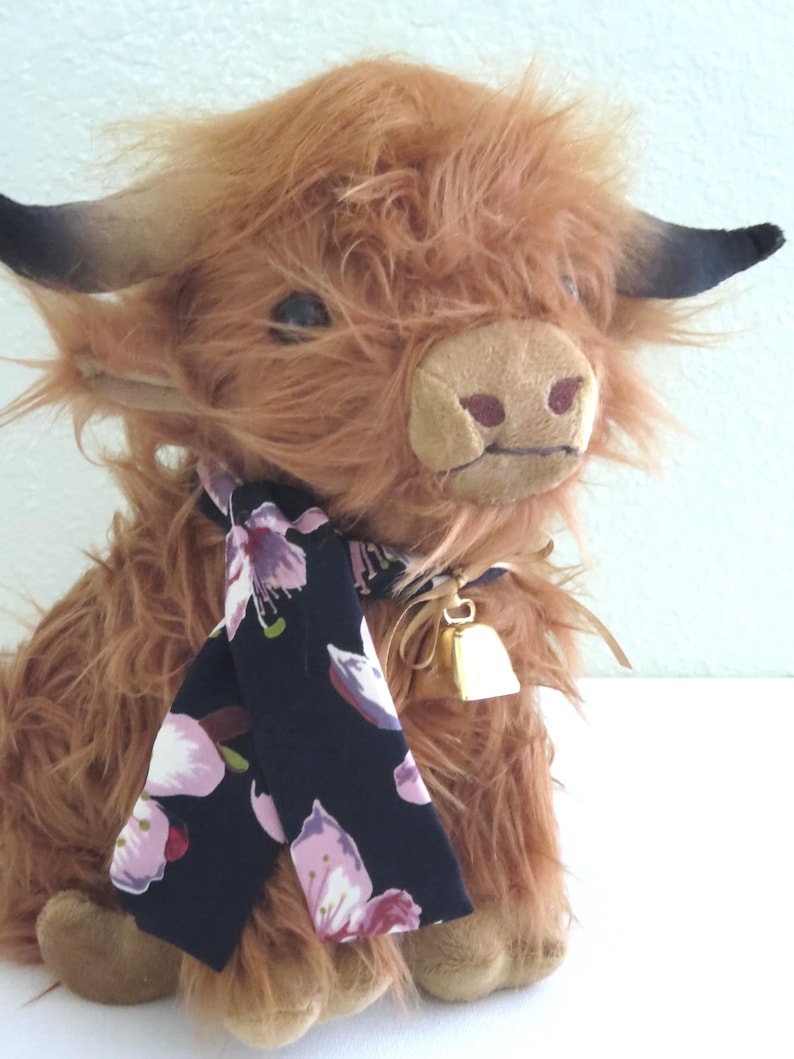 K&ouml;nnte beinhalten: Ein Pl&uuml;sch-Highland-Kuh mit zotteligem braunem Fell, schwarzen H&ouml;rnern und einer beigen Schnauze. Sie tr&auml;gt einen schwarzen Schal mit rosa Blumenmustern und eine kleine goldene Glocke. Das Spielzeug ist einer Highland-Kuh nachempfunden.