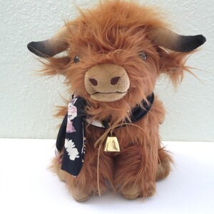 K&ouml;nnte beinhalten: Ein Pl&uuml;schtier, das eine Highland-Kuh darstellt, mit zotteligem braunem Fell, schwarzen H&ouml;rnern und einem schwarzen Schal mit Blumenmuster. Es hat eine kleine goldene Glocke um den Hals. Das Spielzeug steht auf einer wei&szlig;en Oberfl&auml;che.